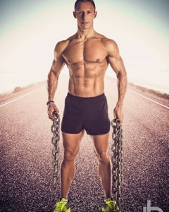 Portrait of Anthony Forsyth: a muscular caucasian man wearing only dark shorts and yellow trainers stands on a sealed road. Hands by his sides he holds two lengths of heavy steel chain.