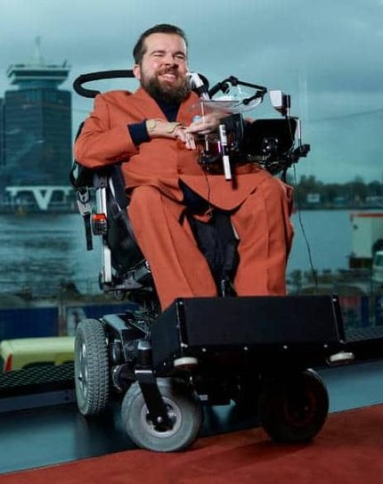 portrait picture of Joost Joseph Nauta, a white male with a beard in an orange suit in a powered wheelchair.