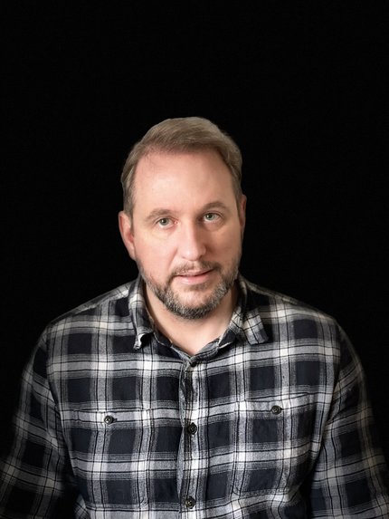 Portrait of James R. Bateman (James Bateman): a man wearing a black and white striped shirt photographed with a dark background. He has a beard and parted brown hair