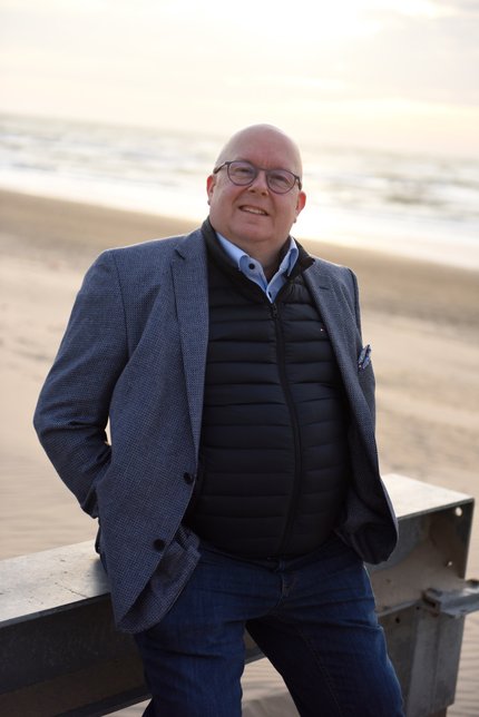 Portrait of Marco Houwen, leaning against a metal support, standing on the beach looking interested.