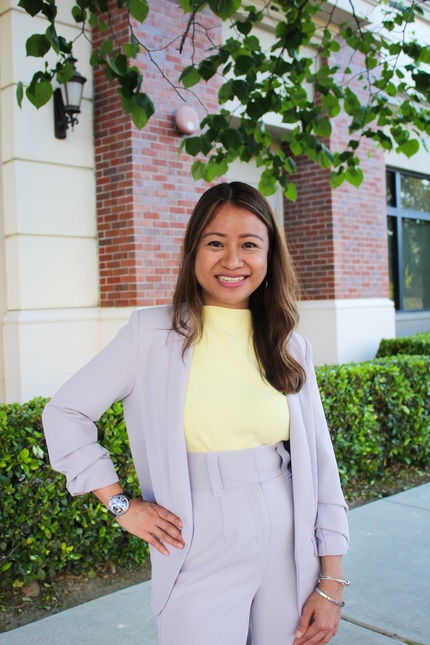 Portrait of Joyce Talag: a slender Asian woman wearing a bright blue top, and a pair of white pants and white high-heeled shoes. She is partly seated on a stool&mdash;one leg stretched and the other rests on the stool&rsquo;s step.