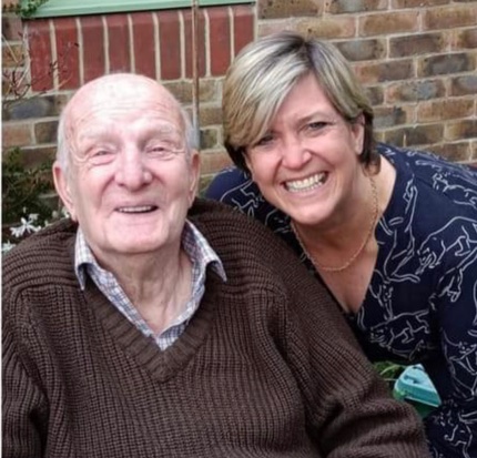 Bald elderly gentleman with his middle aged daughter looking over his shoulder. He’s wearing a brown sweater and she’s got short styled hair and wearing a blue blouse with jaguar print.  Both are smiling