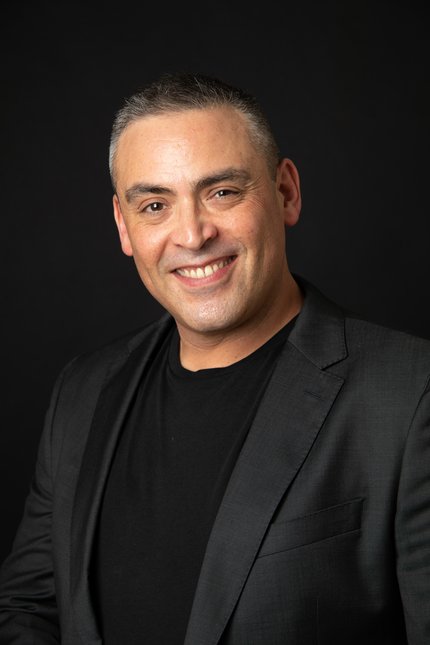 Handsome and strong looking man with olive skin with dark hair, smiling at the camera. He is wearing a dark grey suit, with a black T-Shirt underneath and is stood in front of a black backdrop.