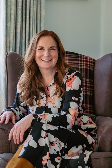 Picture of Mary Ann McGowan: a white Irish female with red hair wearing a dress. She is sitting on an armchair smiling and relaxed.