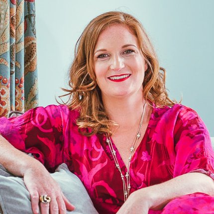 Picture of Mary Ann McGowan: a white Irish female with red hair wearing a pink dress. She is sitting on a couch smiling and relaxed.