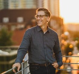 Portrait of Pete Canalichio: a medium build Caucasian man wearing dark denim jeans and a charcoal gray long sleeve shirt. He is standing on the top of a parking deck with his right hand resting on the top rail of a fence surrounding the deck.