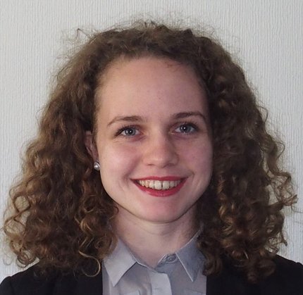 Headshot of a smiling woman with shoulder-length, dark blonde curly hair. She is wearing a grey blouse and black blazer.