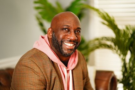 Portrait of Steve Worthy: a relaxed man wearing casual wear of a pink hooded sweatshirt with a brown blazer with corresponding colors. Steve's expression has a soft and relaxed smile with tropical greenery in the backdrop of the picture.