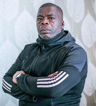 Portait of Duke Harrison-Hunter a black man wearing a black addidas tracksiut top with 3 stripes down the arm with his arms folded, side view